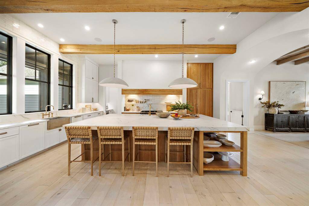 This spacious kitchen features a large island with seating, modern pendant lighting, and a mix of wood and white cabinetry. It has ample natural light from large windows, sleek countertops, and farmhouse sinks, offering a blend of warmth and contemporary design.