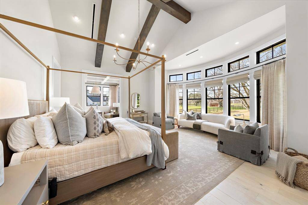 The primary bedroom features high vaulted ceilings with exposed beams and natural light from a wall of large windows overlooking a serene outdoor view. The room includes a comfortable seating area, neutral tones, and elegant window treatments for a relaxing ambiance.