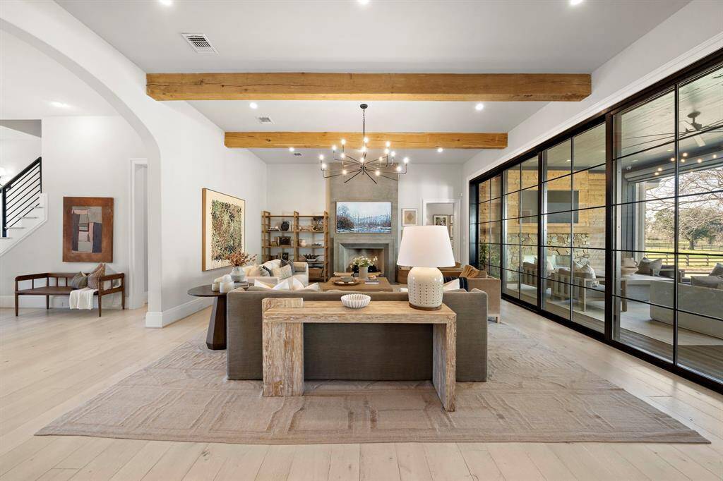 The spacious living room features high ceilings with wooden beams, accordion glass doors, and a cozy fireplace. The open layout is perfect for comfortable living and entertaining.