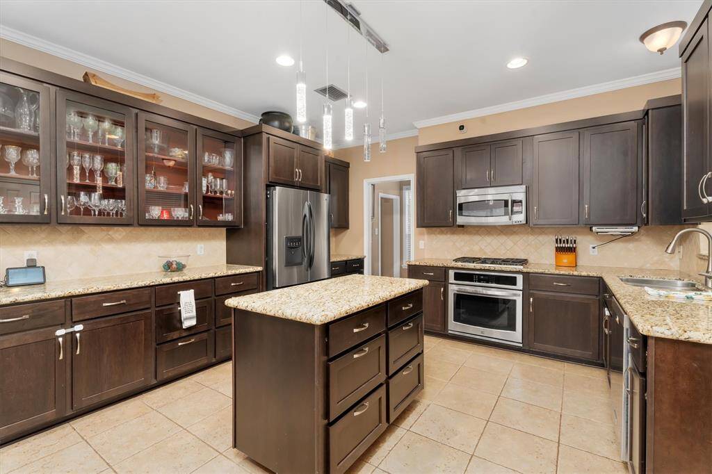 This kitchen features dark wood cabinets, granite countertops, and stainless steel appliances. There's an island with storage, tile flooring, and glass-front cabinets for display. Modern pendant lighting adds a stylish touch.