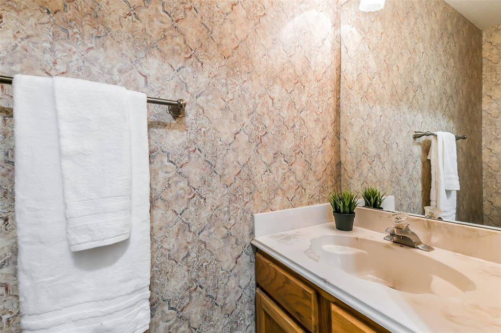 A half bathroom is located under the staircase to service first-floor guests. It offers a cultured marble countertop with a single basin sink, wood cabinetry, and wallpaper.