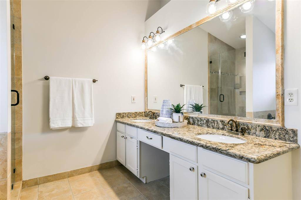 The primary bathroom has been impressively updated to include a lengthy seated vanity with dual undermount sinks, granite countertops, white cabinetry, a framed mirror, stylish lighting, tile floors, a jacuzzi style bathtub, and a frameless glass shower with a tile surround.