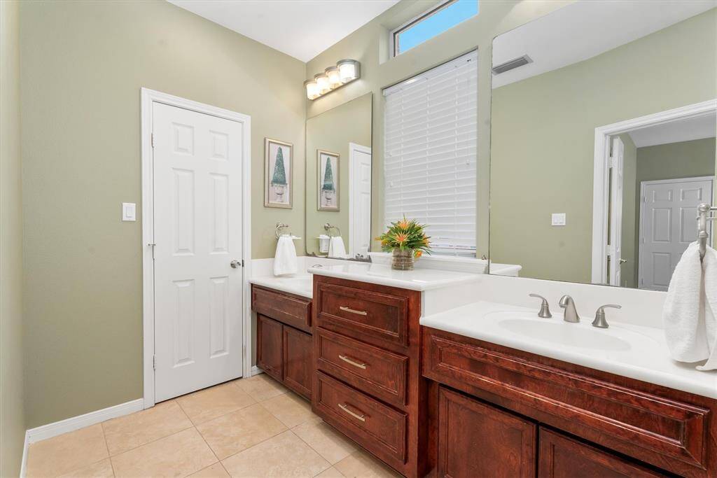 The primary bathroom features a dual-sink vanity with rich wood cabinetry and modern fixtures. It has a large mirror, ample lighting, and an additional window for natural light. The walls are painted a soft green, complementing the neutral tile flooring.