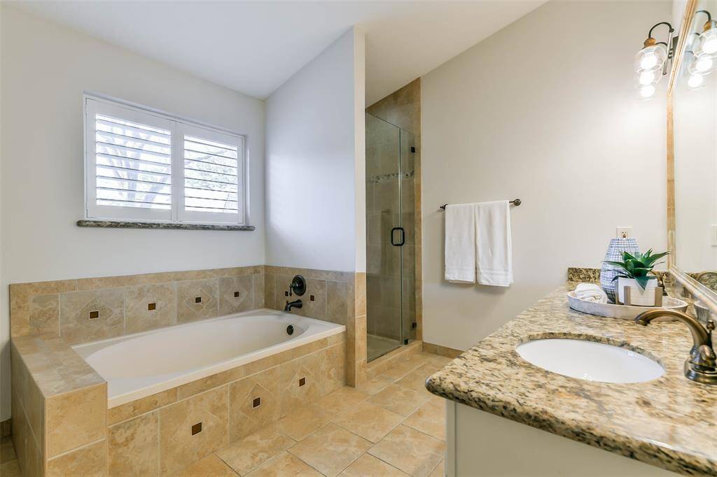 The primary bathroom has been impressively updated to include a lengthy seated vanity with dual undermount sinks, granite countertops, white cabinetry, a framed mirror, stylish lighting, tile floors, a jacuzzi style bathtub, and a frameless glass shower with a tile surround.