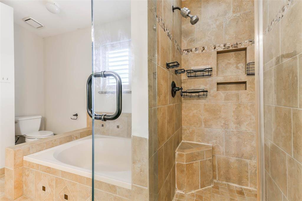 The primary bathroom has been impressively updated to include a lengthy seated vanity with dual undermount sinks, granite countertops, white cabinetry, a framed mirror, stylish lighting, tile floors, a jacuzzi style bathtub, and a frameless glass shower with a tile surround.