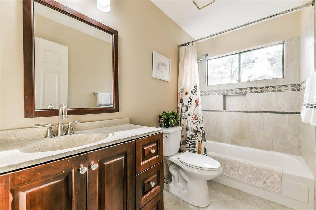This bathroom services the upstairs bedrooms and game room with a single stained wood vanity topped by a manufactured stone countertop and basin sink, a framed mirror, tile floors, and a combination bathtub and shower with a tile surround.