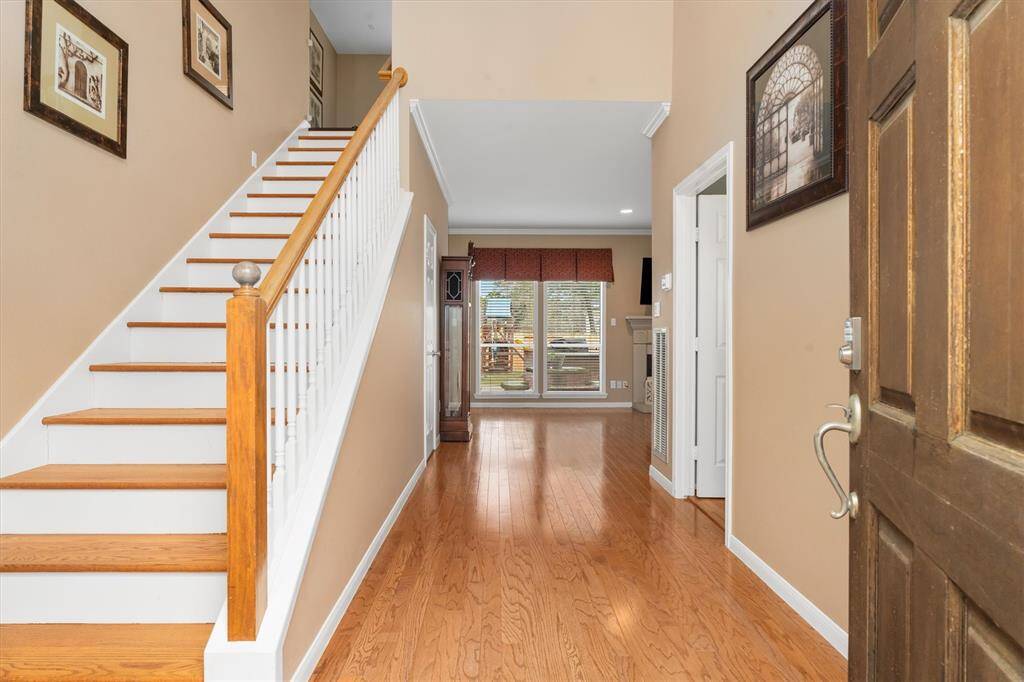 This photo shows a welcoming entryway with warm wood flooring, an open staircase to the left, and a view into a bright living area with large windows. The neutral wall colors and decorative elements create a cozy and inviting atmosphere.