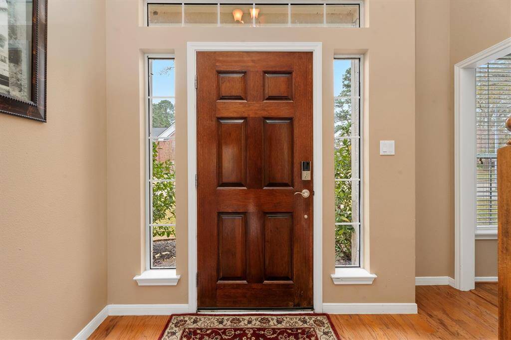 A rich wooden front door, flanked by tall windows allowing natural light to fill the space. The warm-toned walls and wood flooring create an inviting atmosphere.