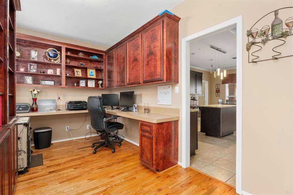 The home office features rich wooden cabinetry and shelving, providing ample storage and display space. It includes a built-in desk area, suitable for work or study. The room opens into a bright kitchen area, creating a seamless flow between spaces.