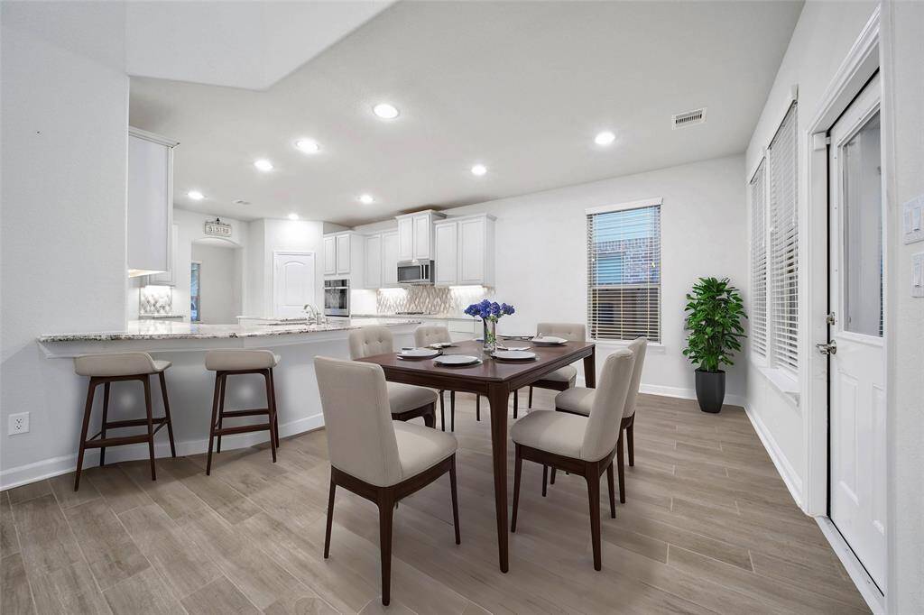 This is a bright, modern open-plan space featuring a dining area and kitchen peninsula with seating. The room has recessed lighting, large windows with blinds, and easy-to-maintain flooring.