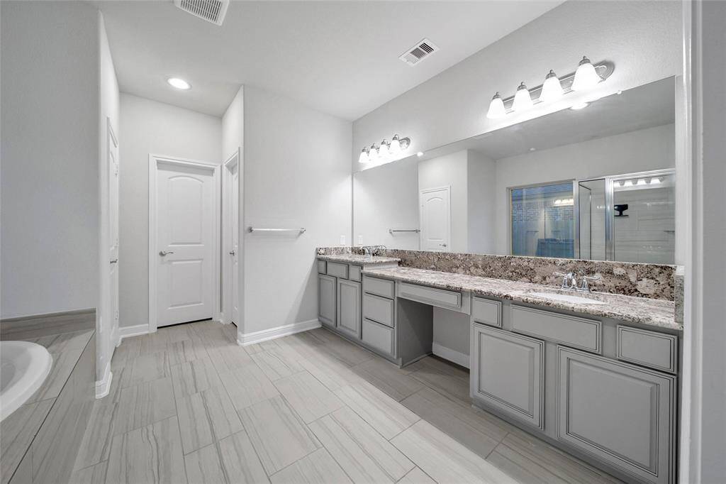 The primary suite bath features a double vanity with granite countertops, ample cabinet space, large mirrors with overhead lighting, and a walk-in shower with glass doors. The room is finished with light wood-like tile flooring, offering a clean and contemporary look.