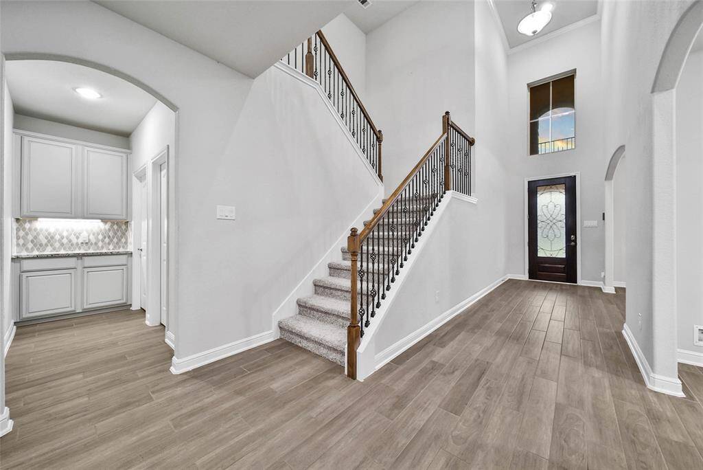 This is a bright, spacious entryway with high ceilings and a grand staircase. There's elegant wood look tile flooring throughout and a tasteful built-in cabinet area to one side. Large windows above the front door allow for natural light to enhance the welcoming atmosphere.