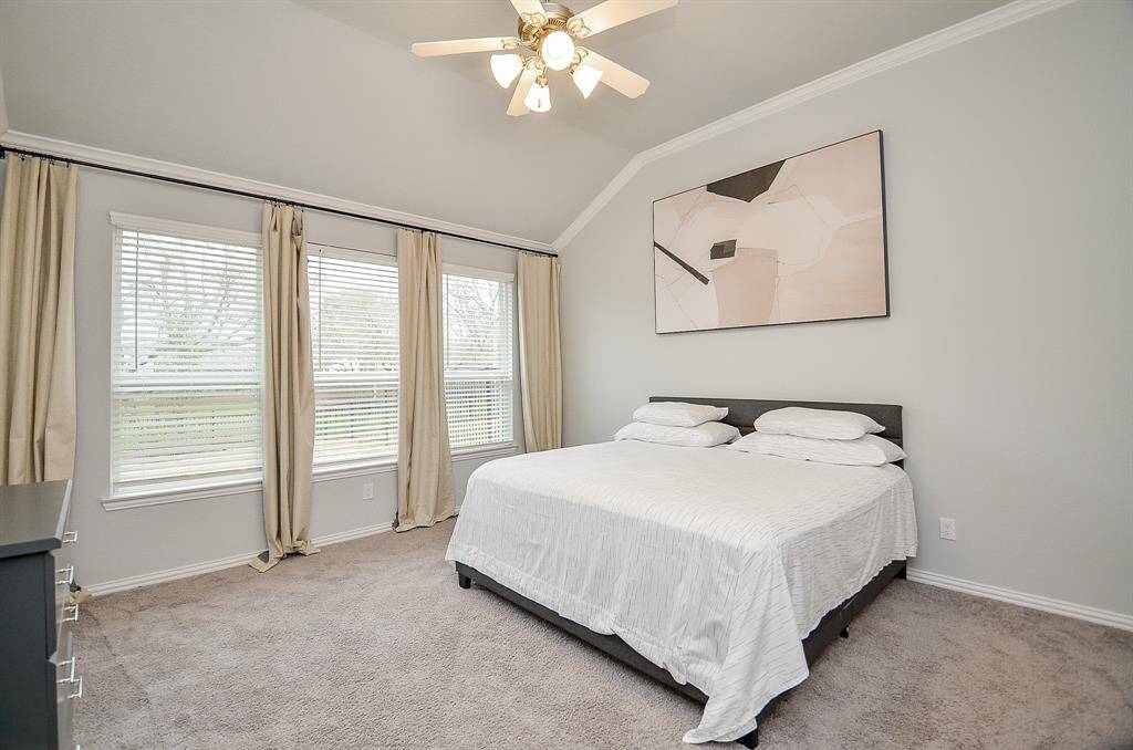 This primary bedroom features a cozy, carpeted floor with Natural light streaming in through three windows with neutral drapes, and a ceiling fan provides comfort. The walls are painted in a soft, calming color.