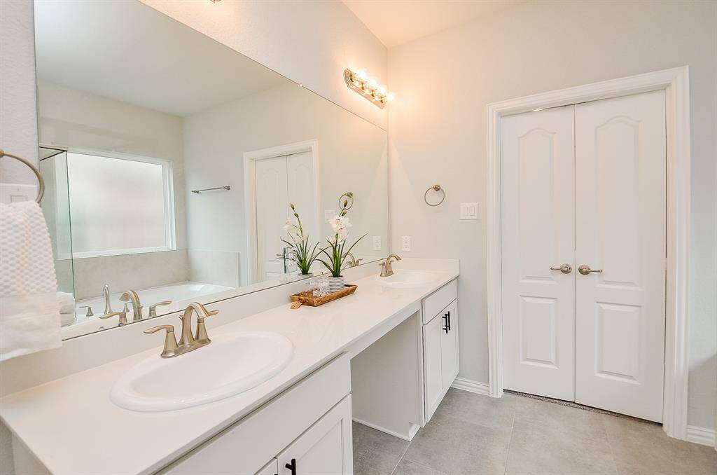 This bathroom features a spacious dual-sink vanity with modern fixtures, a large mirror, and ample lighting. It has neutral tones, a tiled floor, and a separate bathtub area, offering a clean and elegant design.