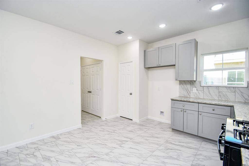A clean, empty kitchen with gray cabinets, marble countertops, and tile flooring. There are recessed lights and a window providing natural light.
