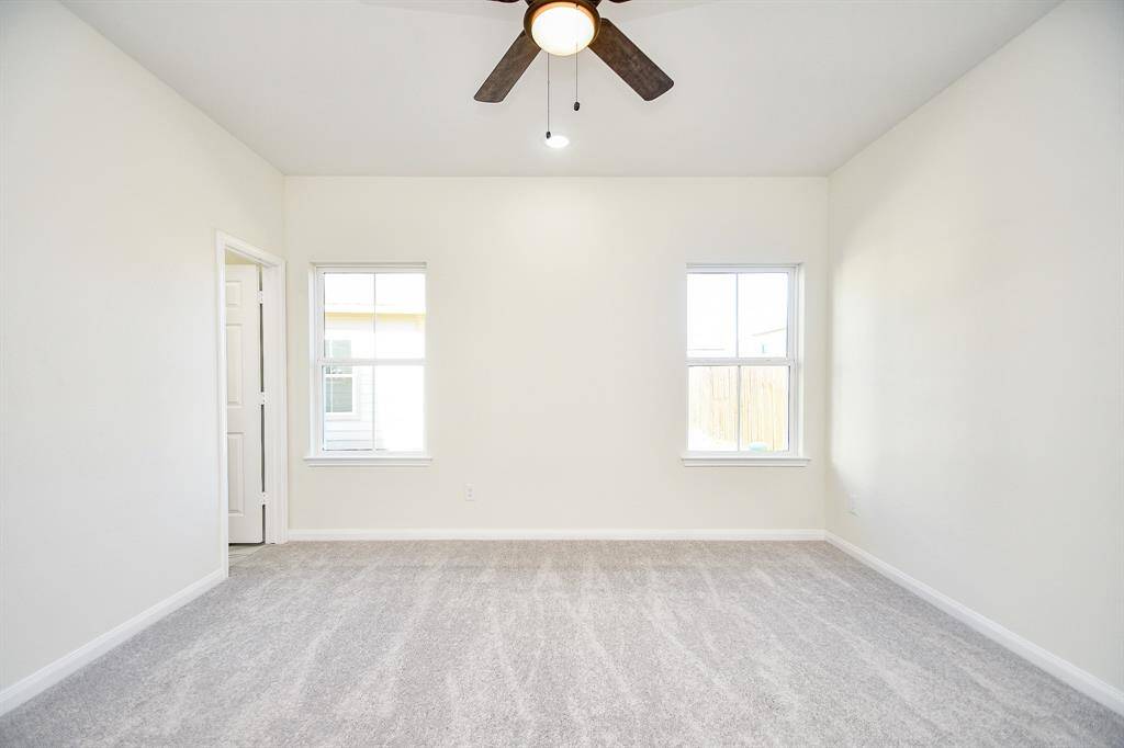 Empty room with gray carpet, white walls, two windows, a ceiling fan, and an open door. Bright and clean space, possibly in a house.
