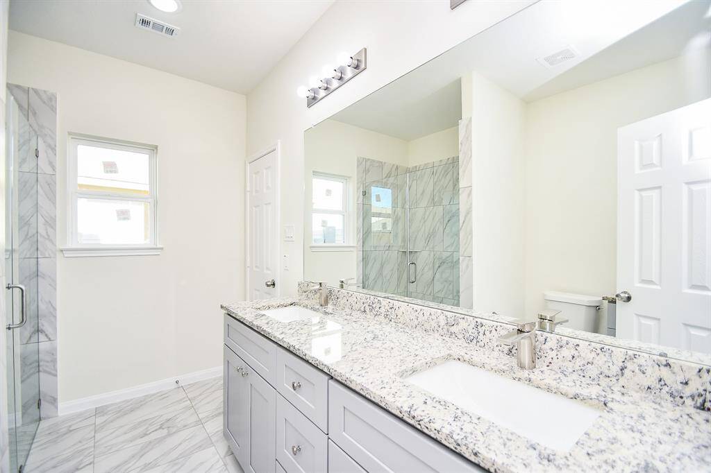 A modern bathroom with dual sinks, granite countertops, large mirror, and a glass-enclosed shower with marble tiles. Bright and clean interior.