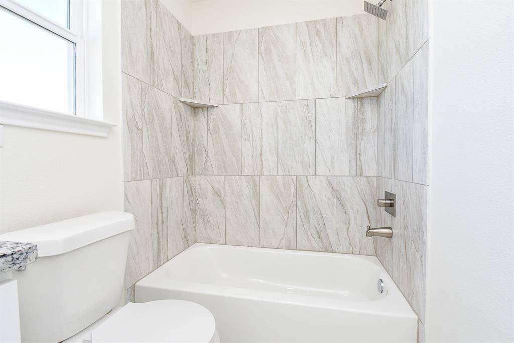 Modern bathroom with a white bathtub, marble-style tiles, built-in shelves, and stainless-steel fixtures, beside a toilet with a window above.