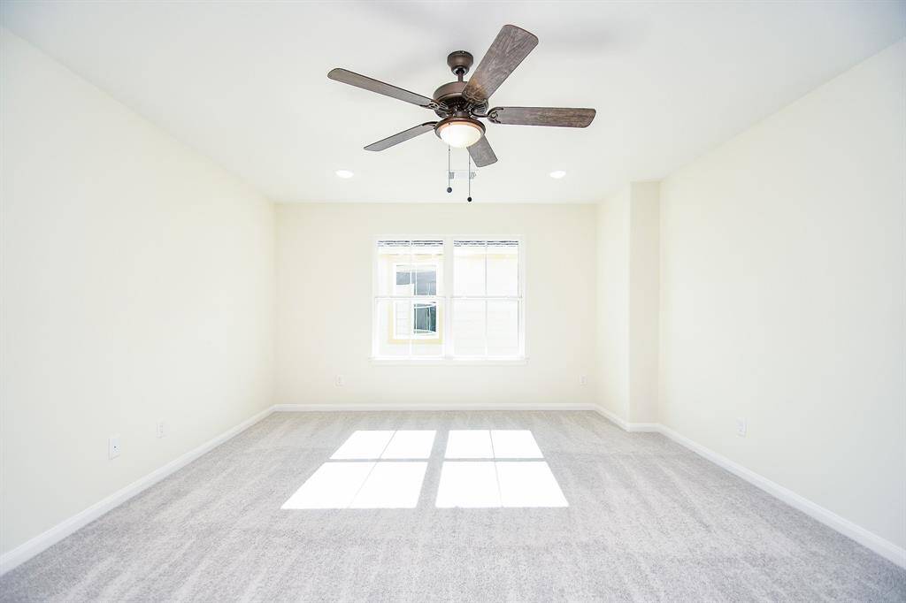 An empty room with beige walls, carpeted floor, a ceiling fan with light, and a single window casting sunlight onto the floor.