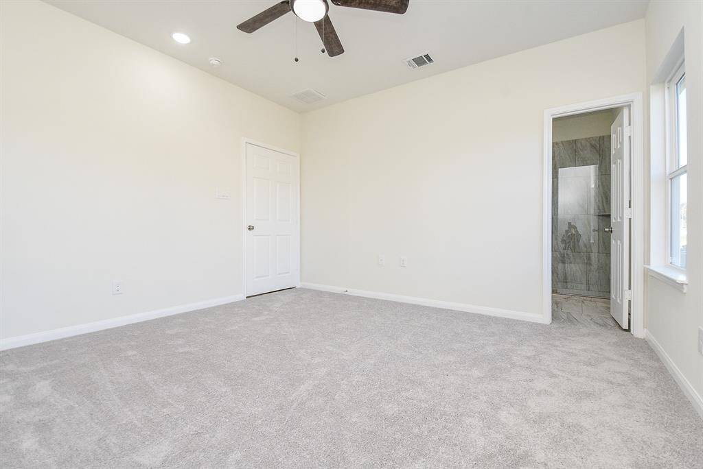 Empty room with carpet, ceiling fan, white walls, a closed door, and open bathroom door showing a tiled shower.