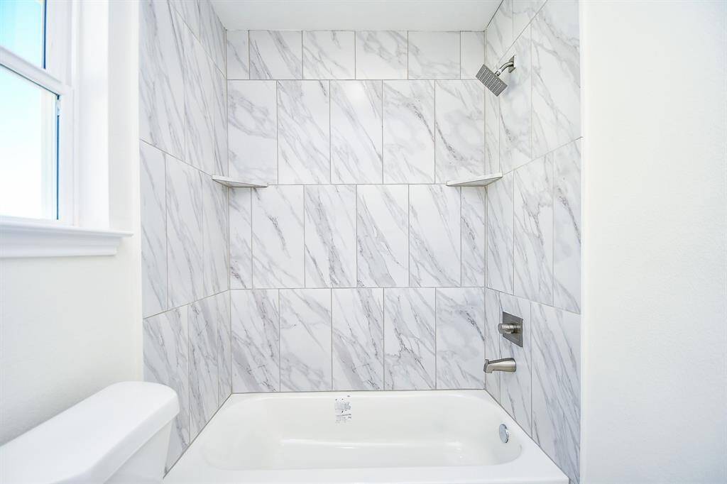 A clean and modern bathroom with a white bathtub and marble-patterned tiles, featuring a built-in shower and natural light from a window.
