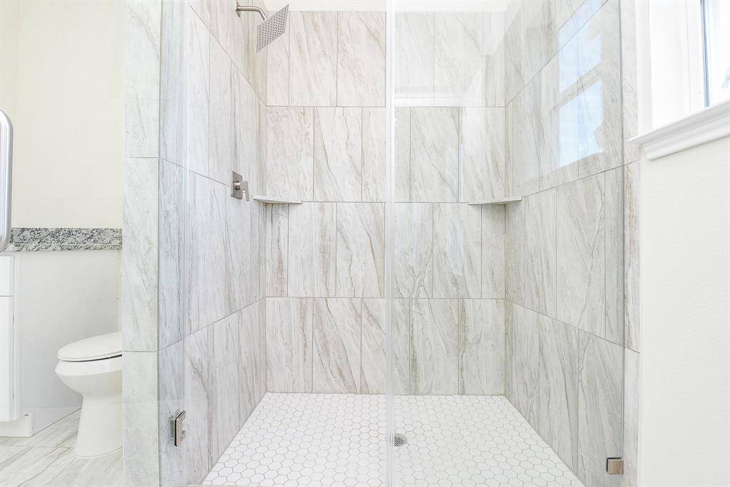 Modern bathroom with marble walls, hexagonal tile floor, glass shower, and white toilet. Bright and clean design with natural light.