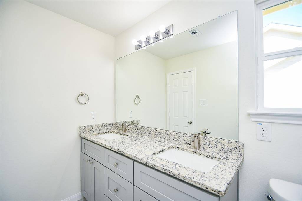 A clean, bright bathroom with a granite countertop, undermount sink, large mirror, and vanity lights. A closed door and toilet are also visible.