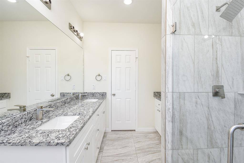 Modern bathroom with double vanity, granite countertops, two mirrors, white cabinetry, and a walk-in shower with marble tiles.