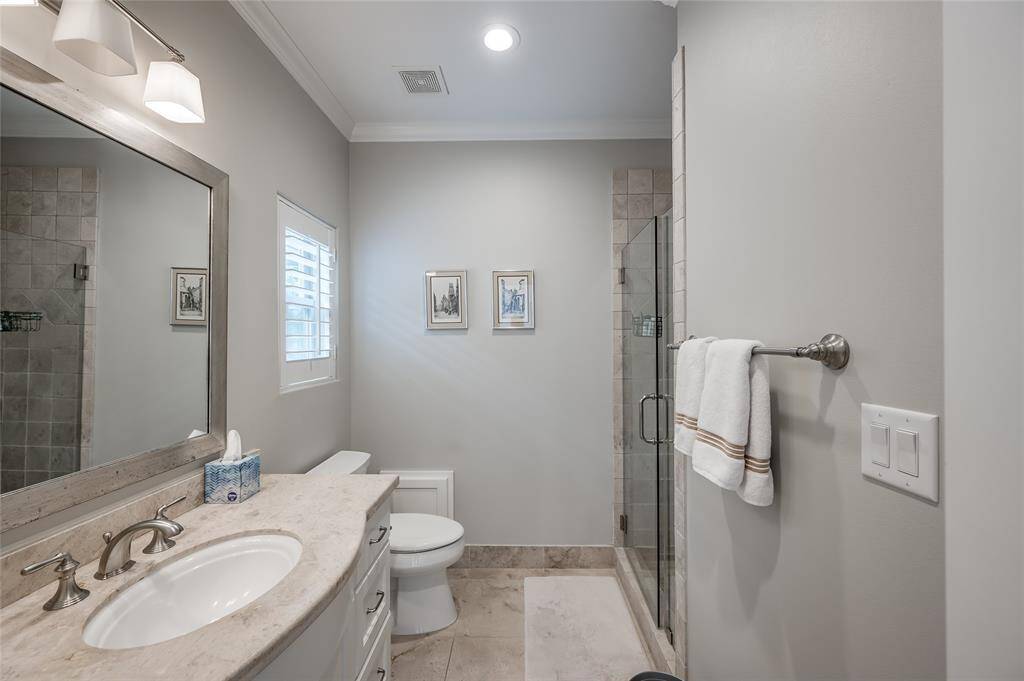 En-suite bath with tile flooring, walk-in shower with tile surround, stone countertops with cabinetry below, and window with plantation shutters