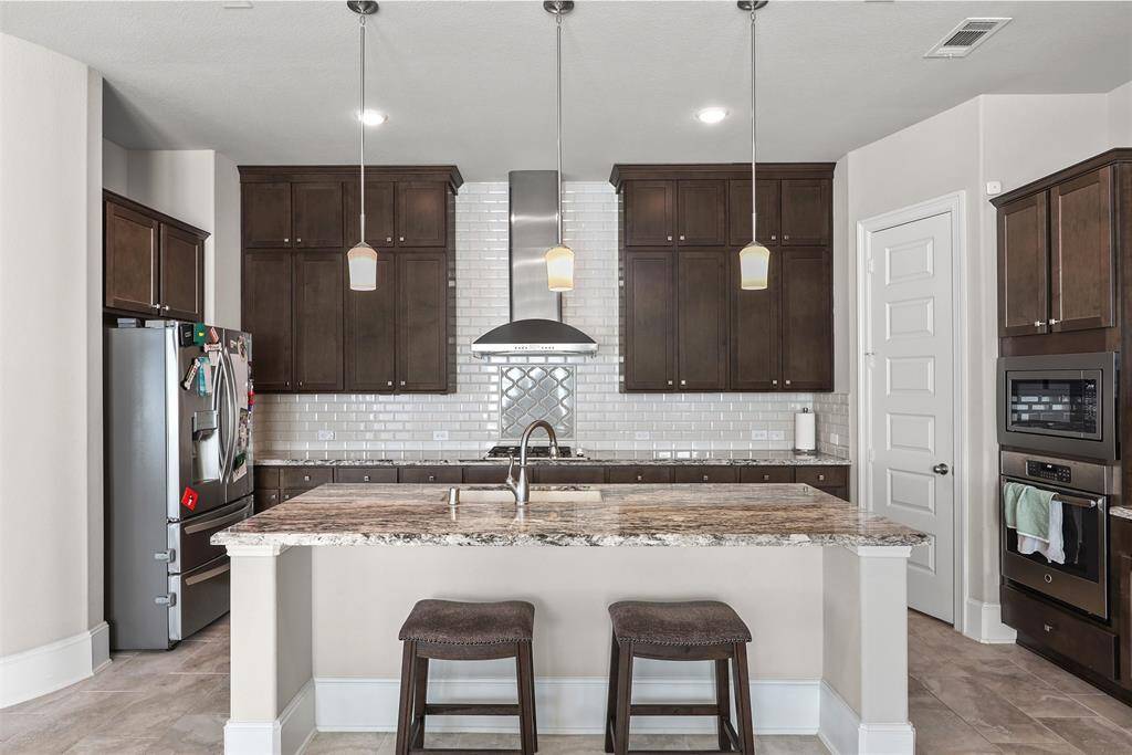 Modern kitchen with dark wood cabinets, stainless steel appliances, and a central island with beveled edge granite countertops. Features pendant lighting, a tile backsplash, extended height cabinets and built-in oven. Ideal for cooking and entertaining.