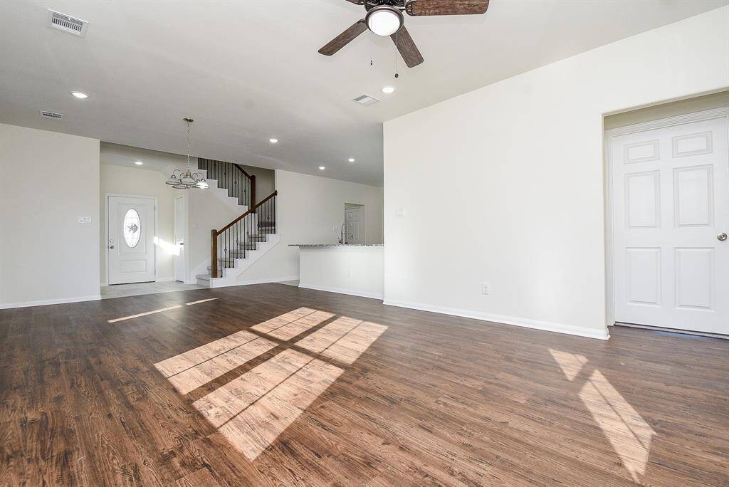 Empty living room with hardwood floors, white walls, a staircase, ceiling fan, and sunlight casting shadows on the floor.