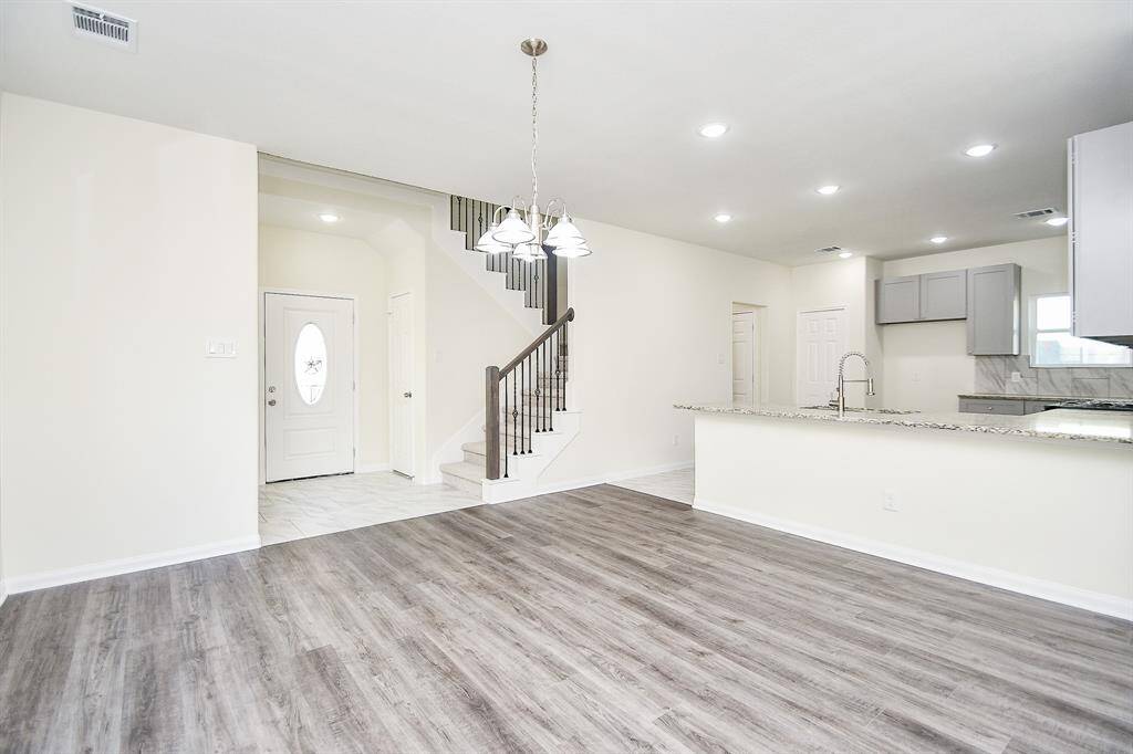 Modern empty interior with a staircase, kitchen area, and pendant lighting; featuring wood flooring and neutral colors.