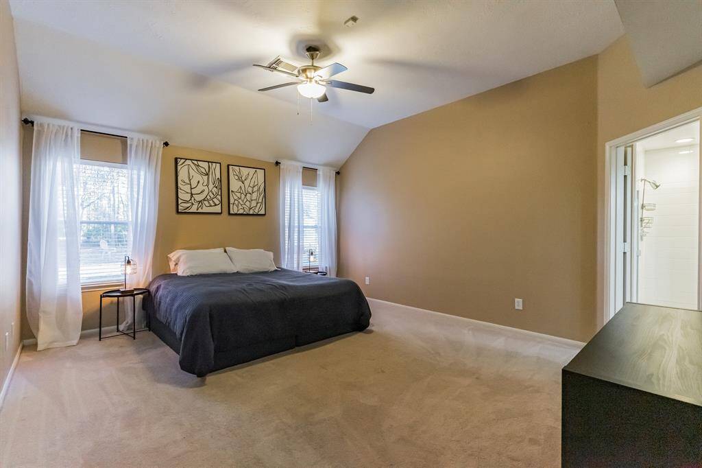 This primary bedroom features neutral walls, a high ceiling with a fan, and ample natural light from two large windows