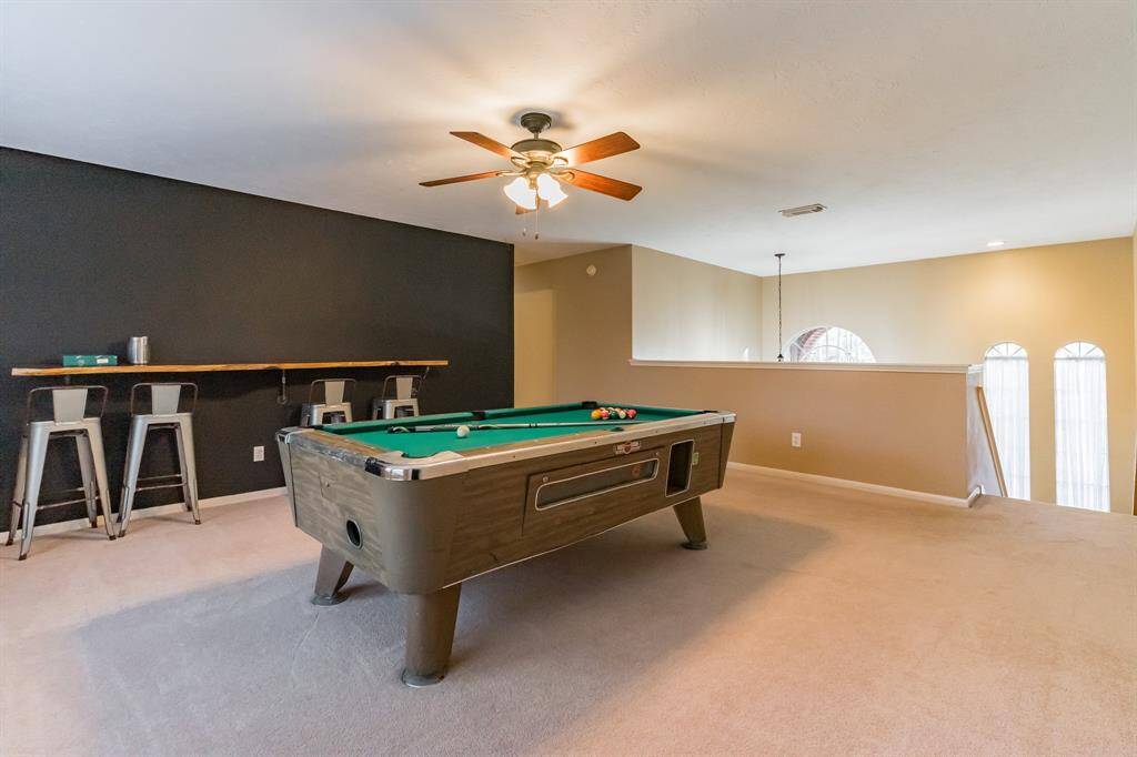 This spacious game room features a pool table, dartboard, and a bar-style seating area. It's well-lit with natural light from two large windows and has a ceiling fan. The room is carpeted and offers a cozy atmosphere for entertainment.