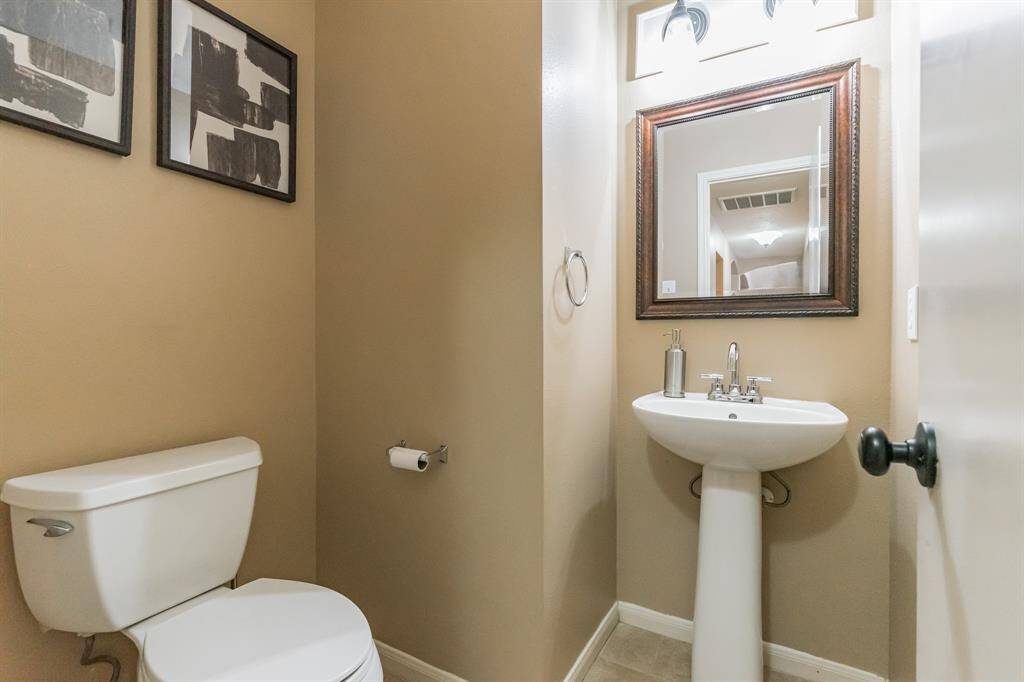 This well-lit bathroom featuring a pedestal sink and a toilet. It has neutral color walls and a framed mirror above the sink. Modern light fixtures add a touch of elegance.