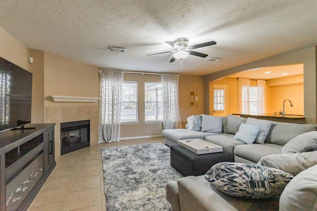 This inviting living room features a cozy fireplace, and a ceiling fan. It has natural light from multiple windows, an open layout to the kitchen, and a modern, neutral color scheme.