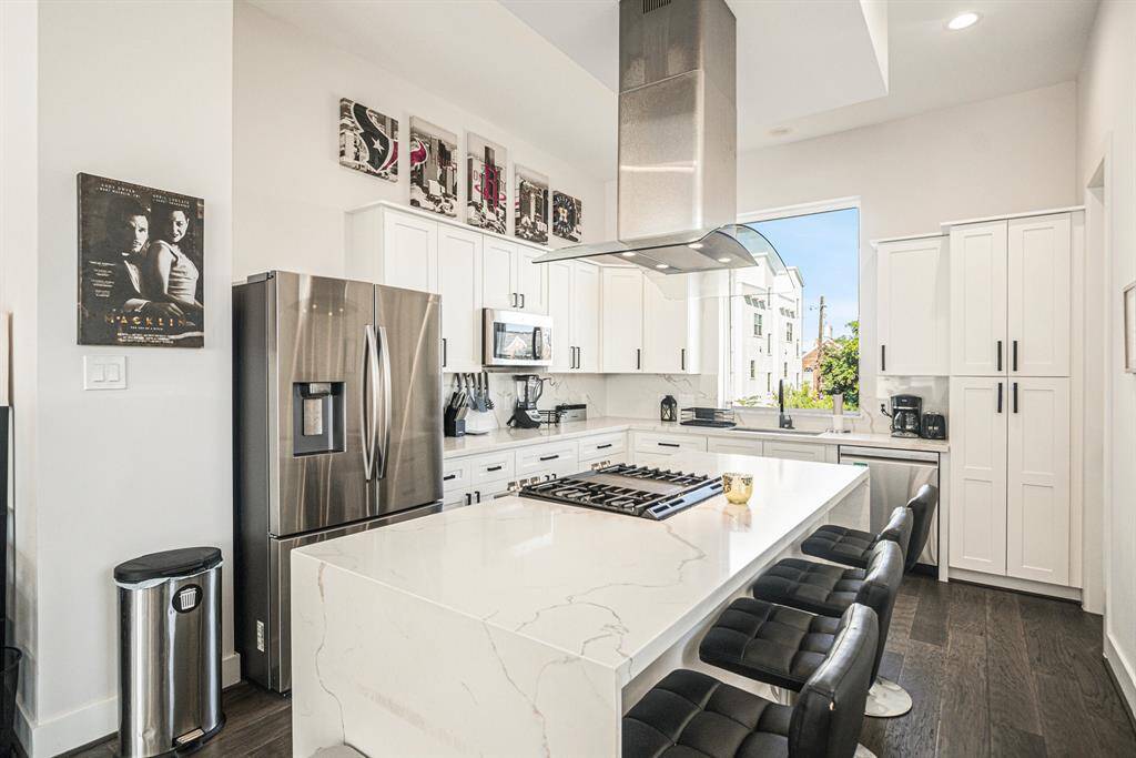 This modern kitchen features sleek stainless steel appliances, a spacious island with a gas cooktop, and elegant white cabinetry. The open layout is accented by dark hardwood floors and large windows, offering ample natural light. Perfect for cooking and entertaining.