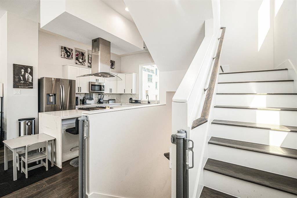 A modern kitchen with stainless steel appliances, white cabinetry, and a sleek range hood. There's a breakfast bar with seating, and adjacent is a staircase with dark wood steps and white risers, leading to a well-lit upper level. The space feels open and contemporary.