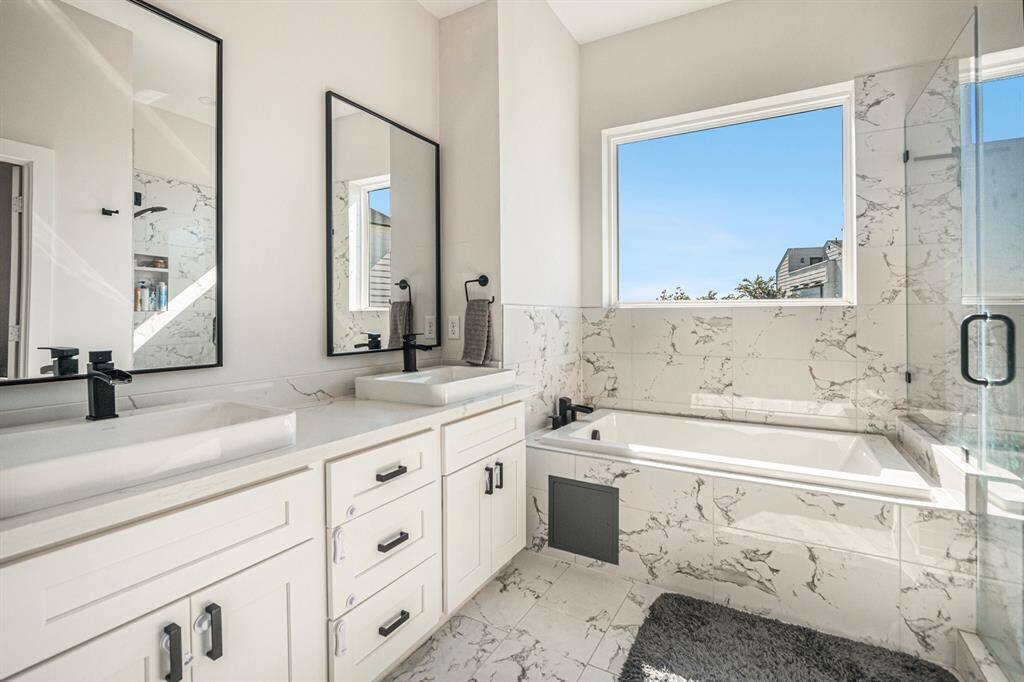 This bathroom features a modern design with dual sinks, elegant black fixtures, and a spacious marble-tiled tub. It offers a bright, airy feel with large windows and a glass-enclosed shower. Perfect for a luxurious and serene atmosphere.