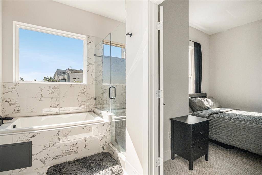 This photo showcases a bright, modern bathroom with a marble-tiled bathtub and glass shower, adjacent to a cozy bedroom.