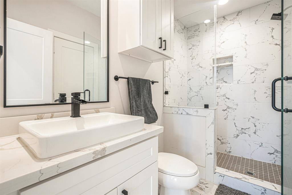 Modern bathroom with a sleek design featuring a white marble vanity and sink. The shower area is enclosed with glass doors and has a stylish, tiled interior. Black fixtures and hardware add a contemporary touch.
