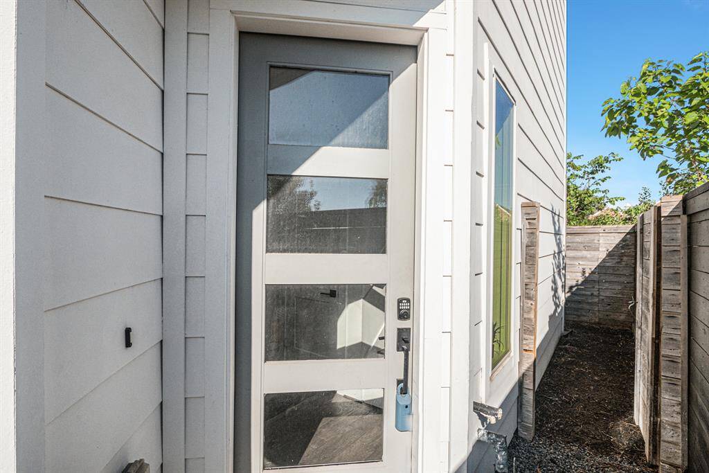 This photo shows a modern side entrance with a glass-paneled door, light-colored siding, and a small fenced pathway leading to the backyard.
