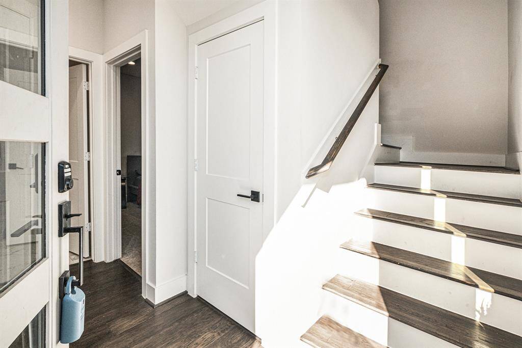 This photo shows a bright entryway with dark wood flooring, a modern white staircase, and two doors leading to adjacent room and bathroom. The space is well-lit, featuring sleek door fixtures and a clean.