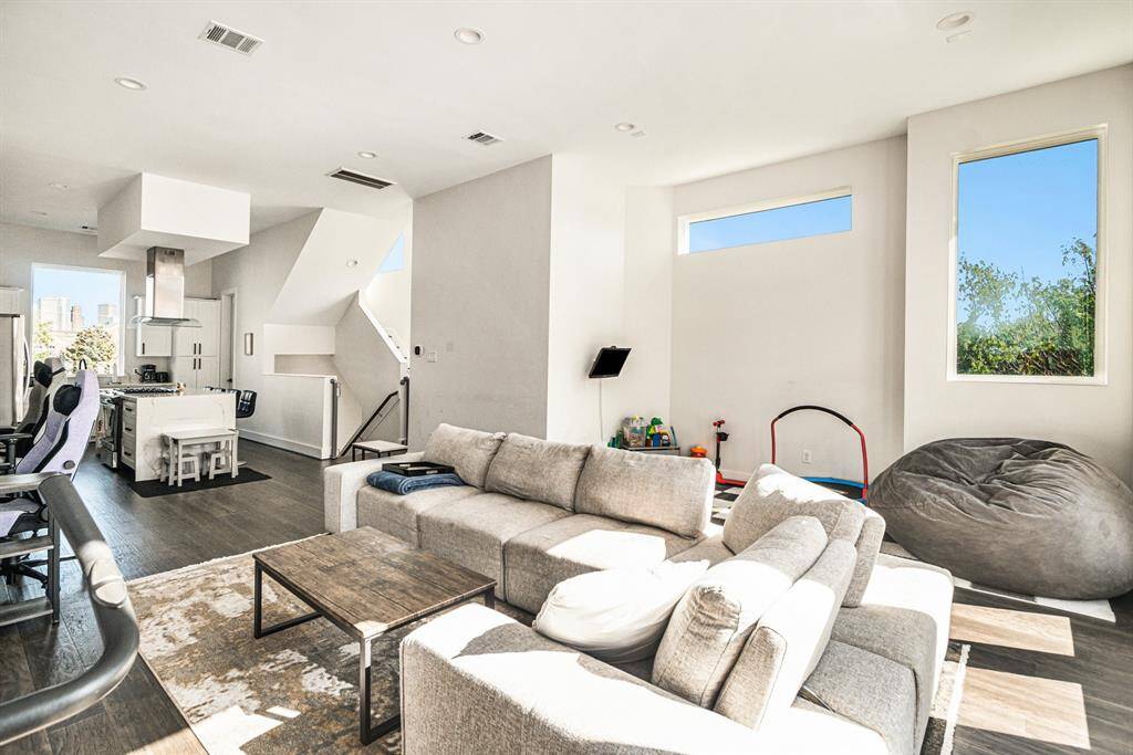 A bright, modern open-concept living space with large windows allowing natural light. The room flows into a sleek kitchen area with a central island, making it ideal for entertaining.