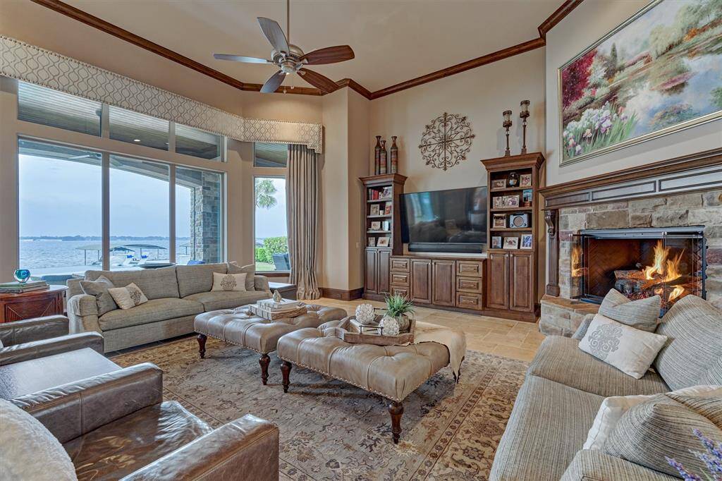 The living room offers tall ceilings, an oversized stone fireplace, custom built-ins and expansive views of the lake. Draperies frame the large windows while a central hidden motorized shade provides privacy.