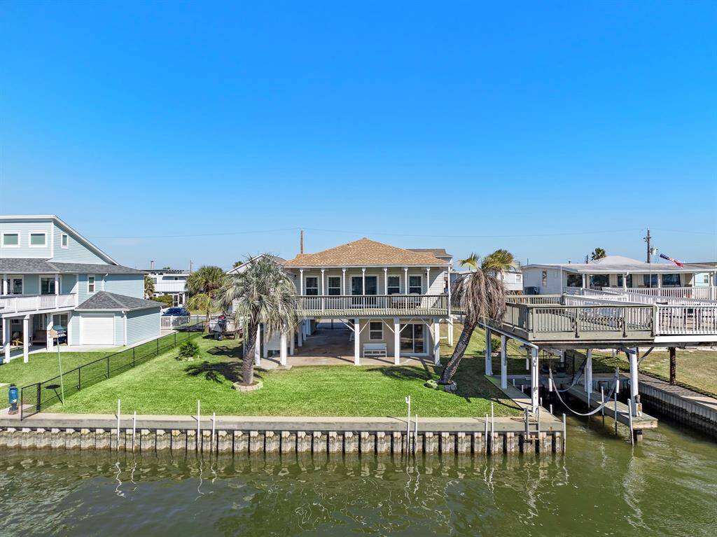 A straight-on view of the home from across the canal. The broad, grassy yard, spacious deck, and covered space below the home are clearly visible, highlighting the generous lot size and prime canal frontage.