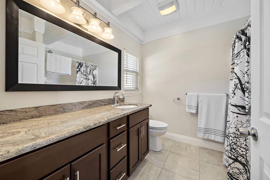 A bright and modern bathroom featuring a granite countertop, rich espresso cabinetry, and a large framed mirror. The design is complete with a tub/shower combo, tile flooring, and classic brushed nickel hardware.