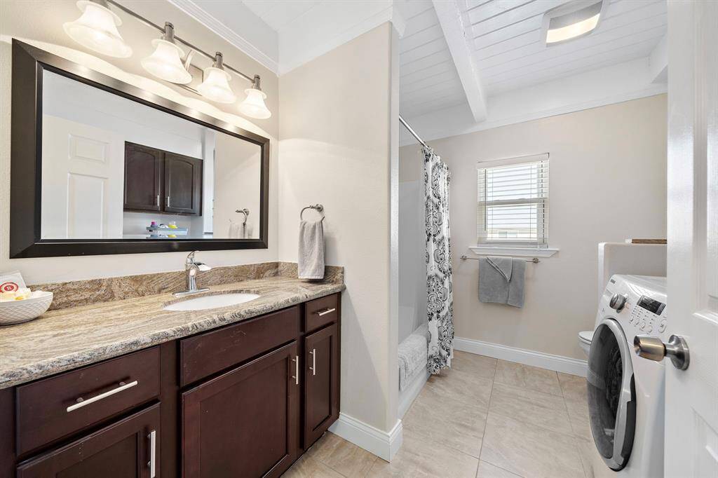 A combined bathroom and laundry space features a sleek granite vanity, stylish cabinetry, and stacked washer/dryer unit for added convenience. The neutral tile flooring and white walls keep the room feeling fresh and spacious.