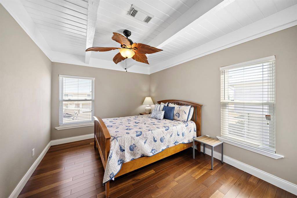 A cozy and charming guest bedroom featuring warm hardwood floors, a nautical-themed bedspread, and large windows that let in plenty of natural light. The neutral tones and tropical ceiling fan give it a coastal retreat feel.