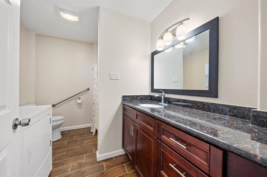A spacious bathroom with granite countertops, rich wood cabinetry, and a wide mirror. The bathroom also features a shower and is equipped with a washer/dryer combo, making it a multi-functional space.
